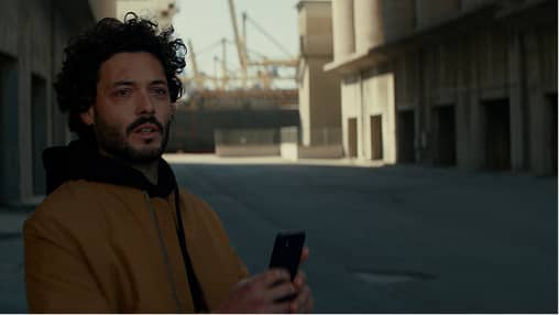 Man with curly hair and beard in brown jacket holding a phone in an industrial outdoor setting.
