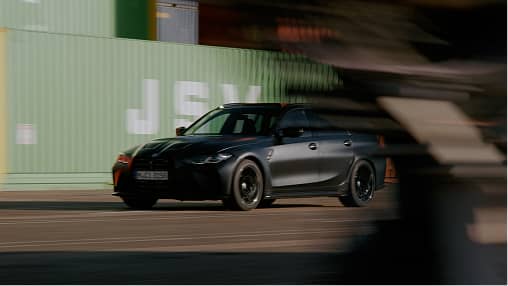 Black car speeding past green shipping containers with motion blur effect.