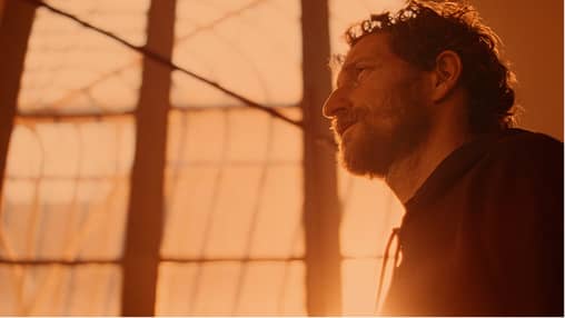 Side profile of a man with curly hair and beard looking through a window with warm sunlight.