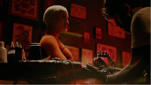 Tattoo artist working on a client's arm in a dimly lit studio with red-toned artwork on walls.