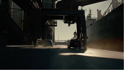 Two cars driving through a dimly lit industrial port area with cranes and a large ship.