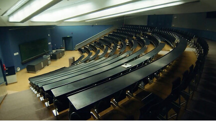 Empty lecture hall with tiered black desks and chairs under bright ceiling lights.