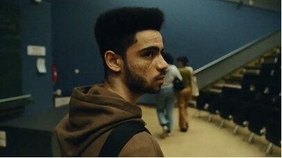 Young man with backpack looks to his right in a dim auditorium with chairs and stairs.