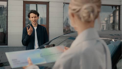 Man holding car keys looking at woman holding a large check by a car outside a building.