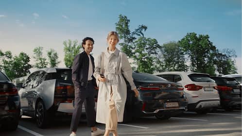 Man and woman walking in a parking lot with cars and trees in the background on a sunny day.