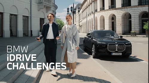 Two people walking on a city sidewalk near a parked black BMW car under a blue sky.