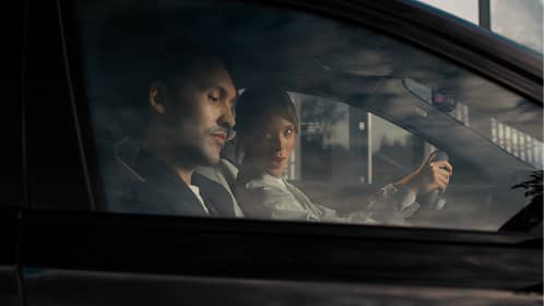 Two people in a car viewed through the passenger window, one driving and the other in the passenger seat.