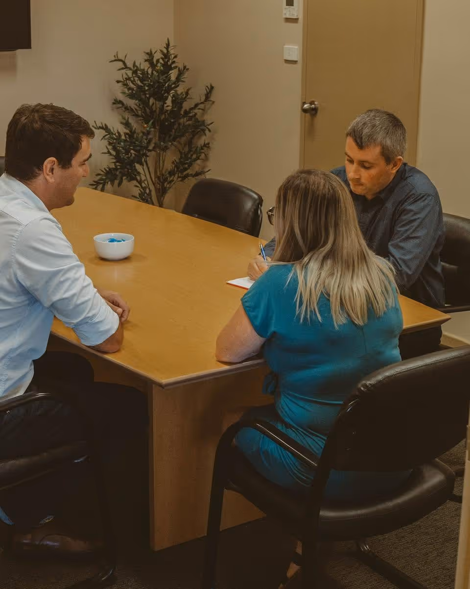 Three people seated around a wooden table in an office, with one person writing in a notebook.