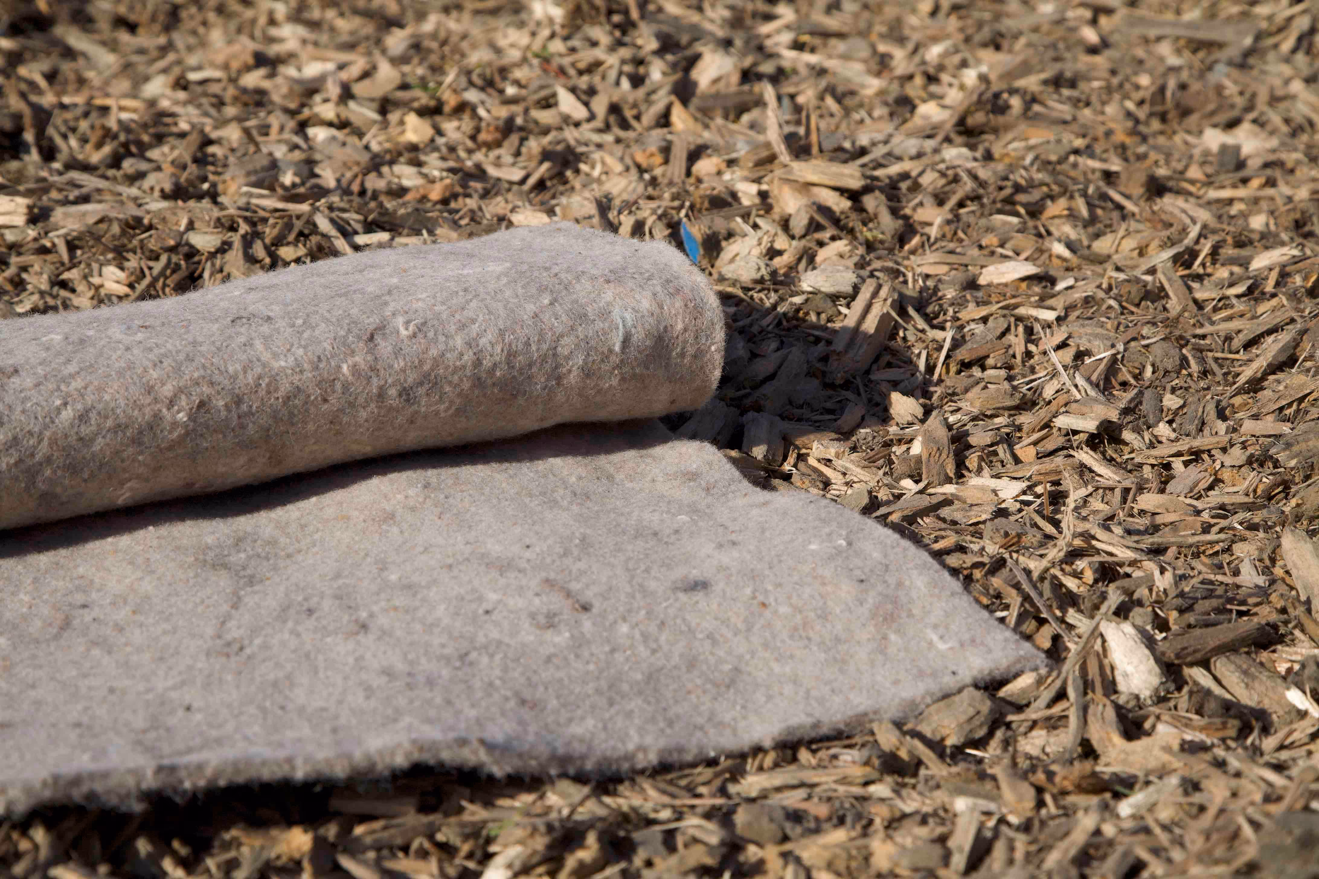 Rolled grey felt mat lying on wood chips outdoors in sunlight.