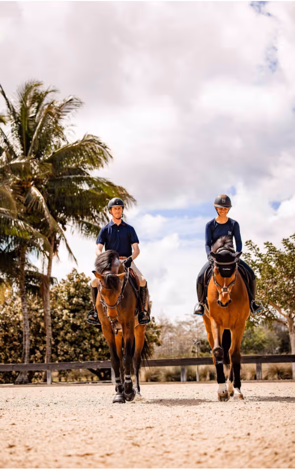 Two riders in helmets on brown horses riding on sandy ground with palm trees and cloudy sky behind.