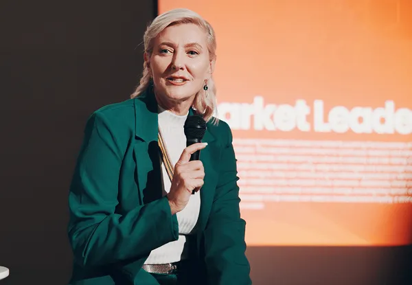 Speaker in a green suit holding a microphone on stage in front of a Market Leaders screen