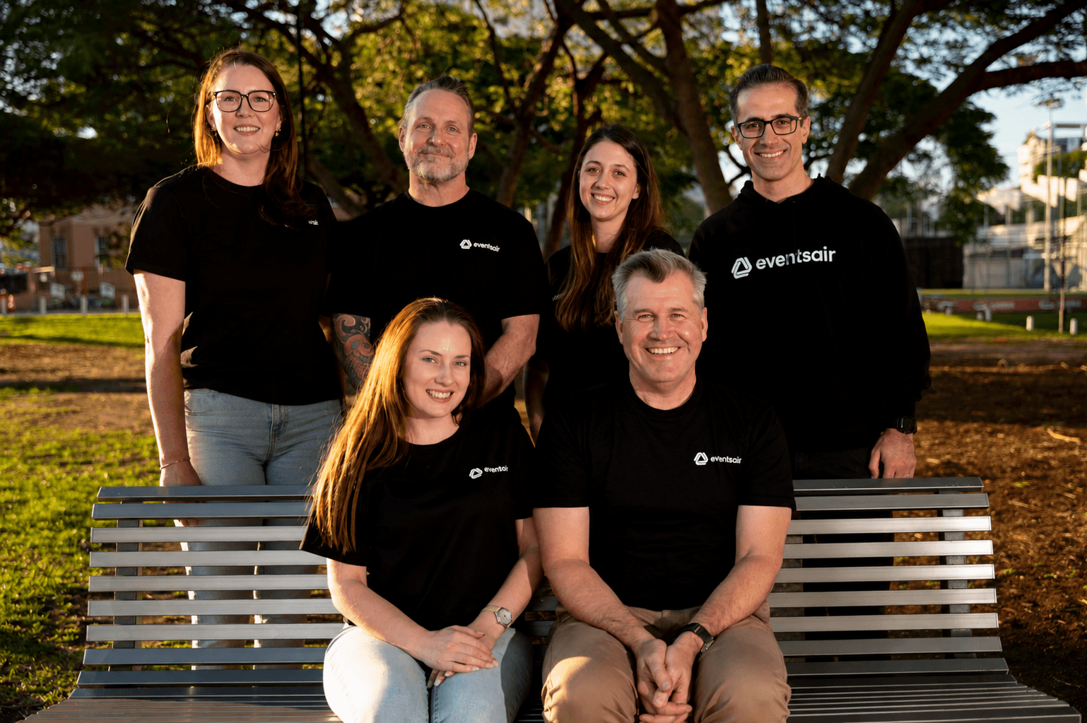 Six EventsAir team members in branded T-shirts posing around a park bench