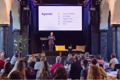 Speaker on stage presenting an agenda slide to a seated event audience