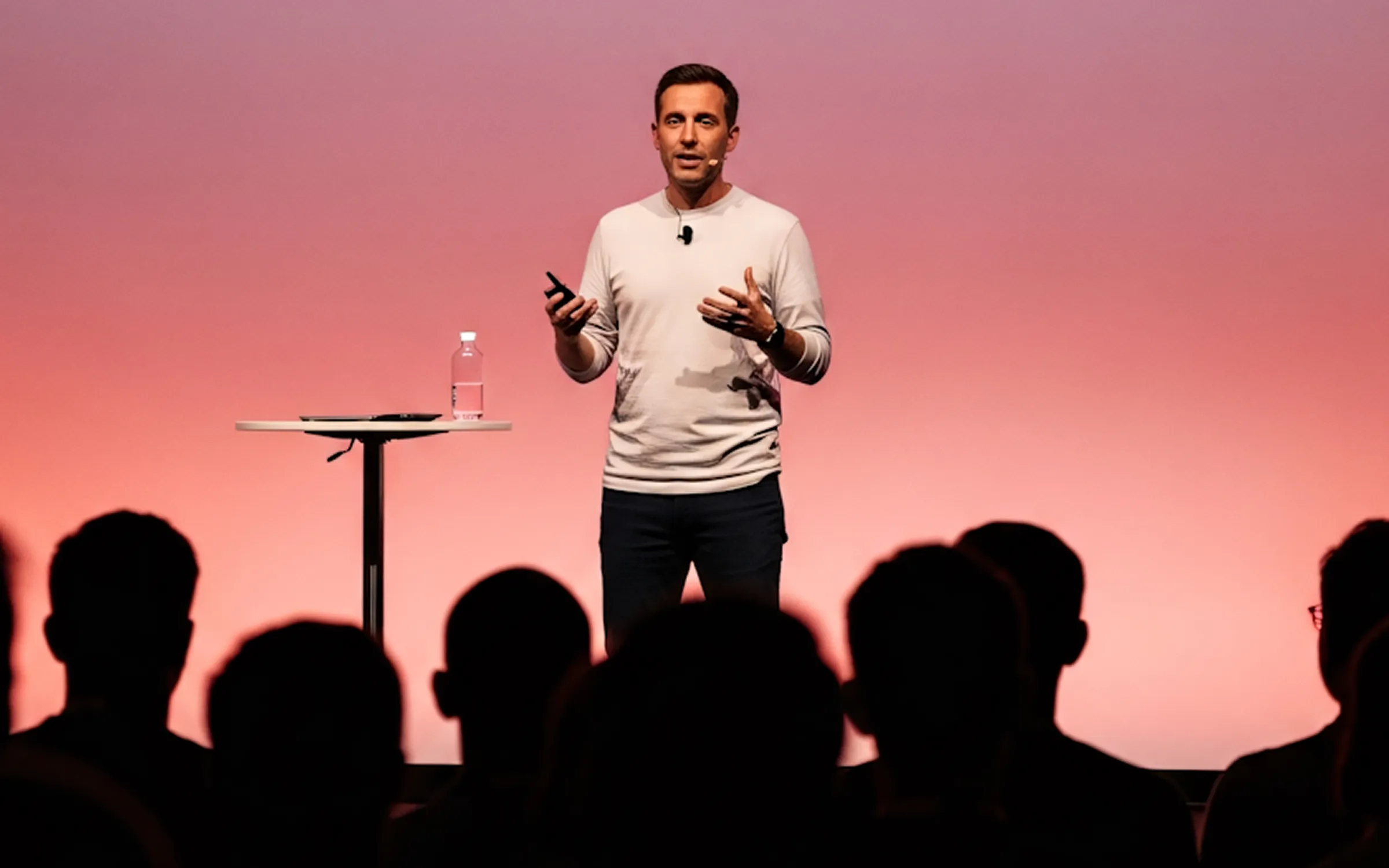 Man giving a presentation on stage with a pink background and an audience in silhouette.