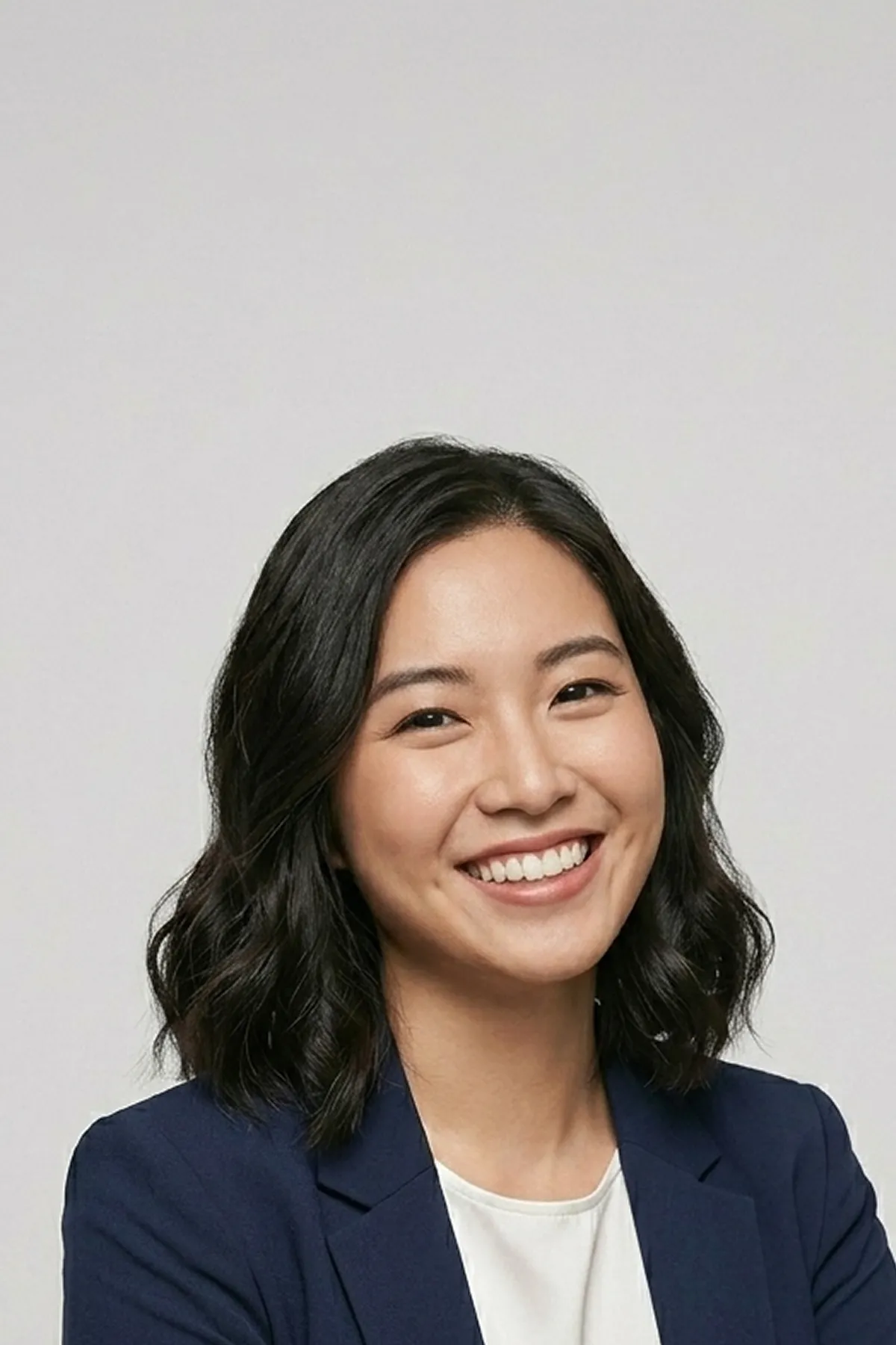 Smiling woman with shoulder-length black hair wearing a navy blazer and white top against gray background.