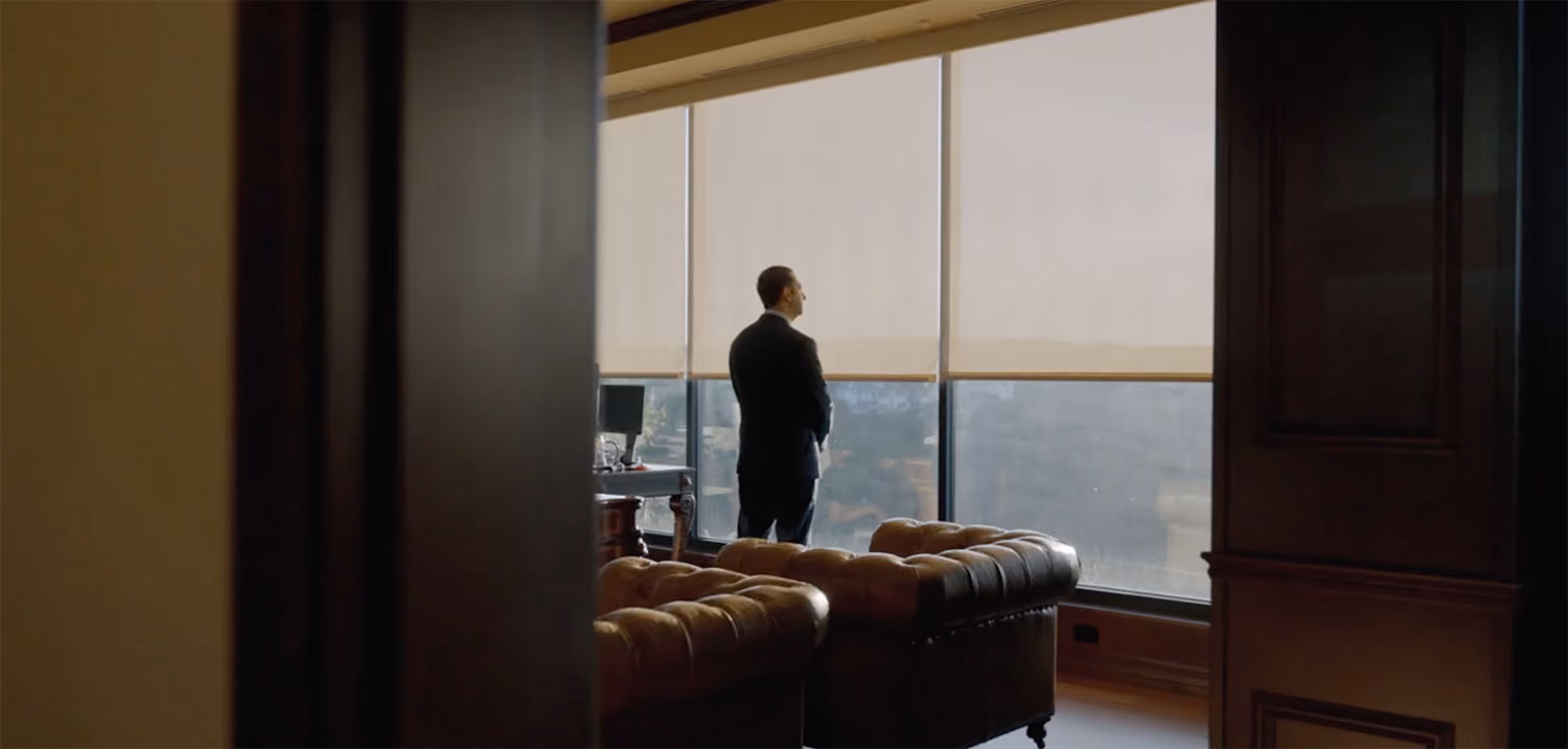 Man in a suit standing in an office looking out large windows with half-drawn blinds.
