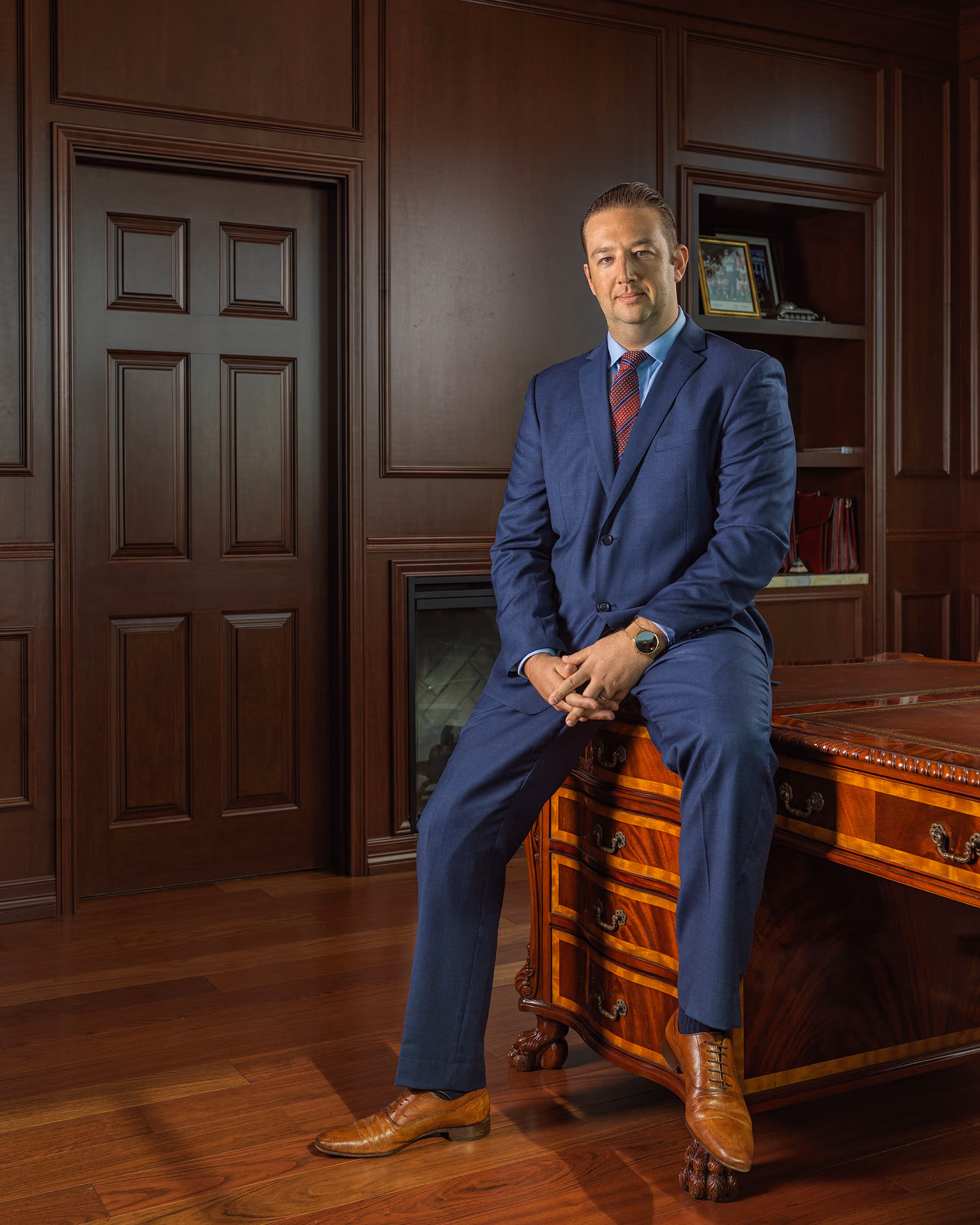 Man in blue suit with red striped tie sitting on an ornate wooden desk in an office.