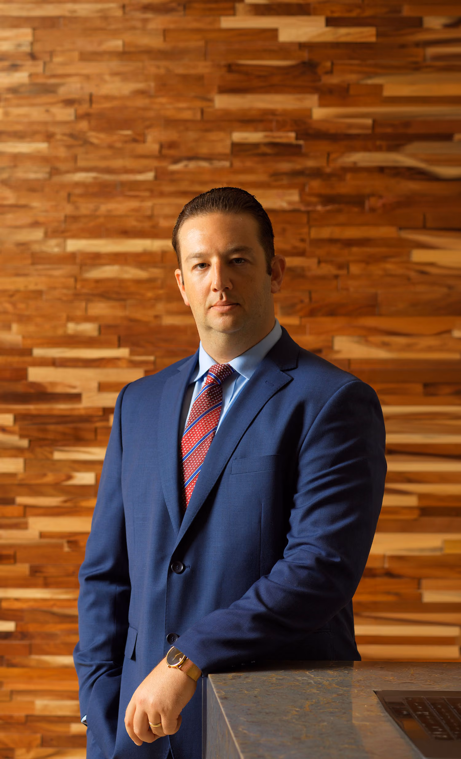 Man in blue suit with red striped tie leans on a stone ledge against a wooden wall.