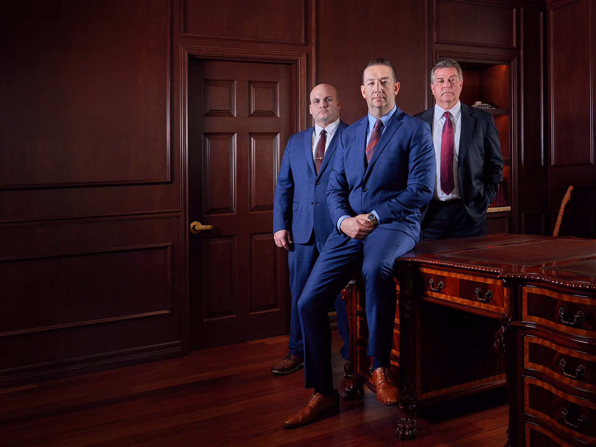 Three men in suits posing in a dark wood-paneled office with a large ornate wooden desk.