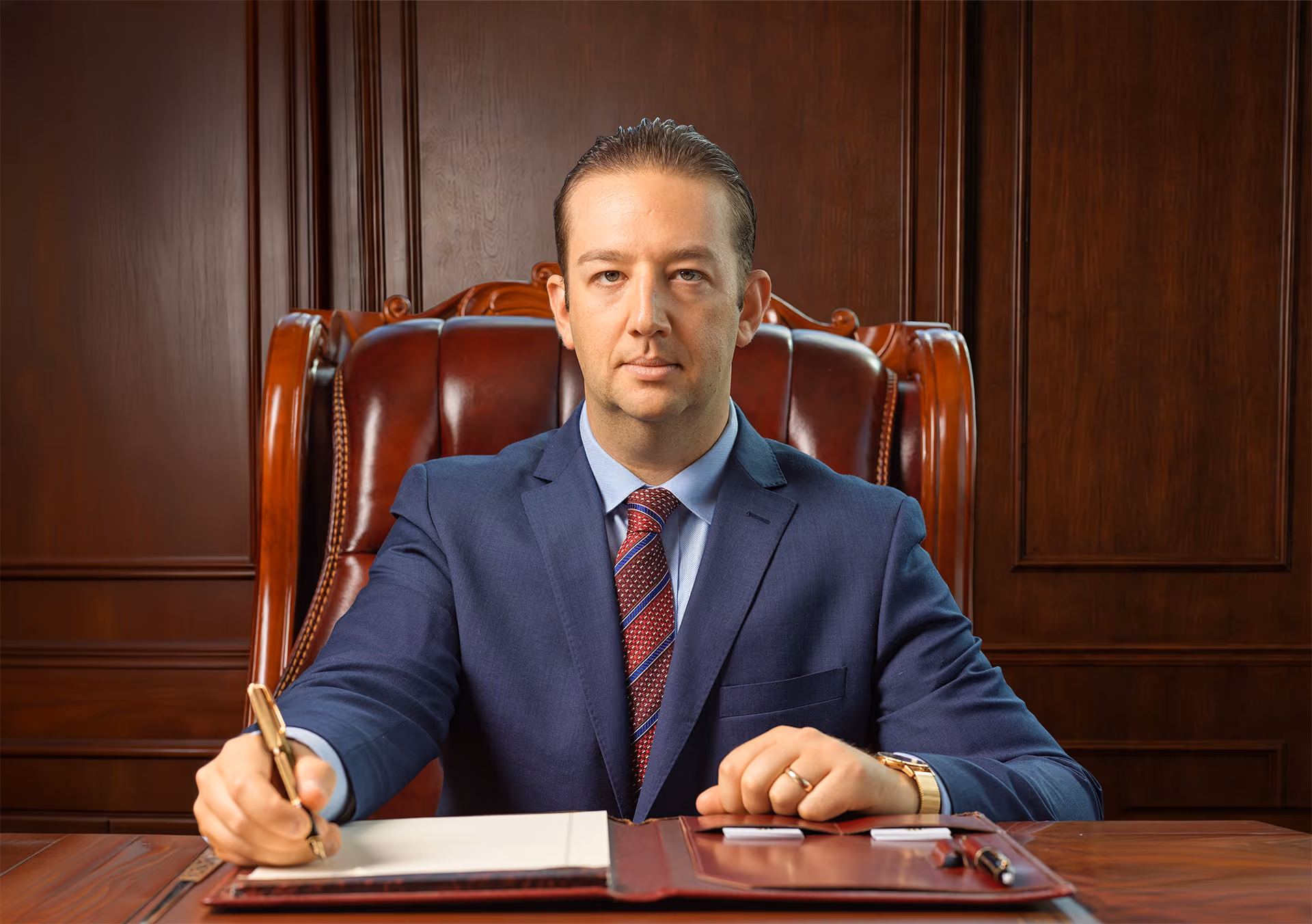 Man in a blue suit and red tie sitting at a desk holding a pen ready to write.