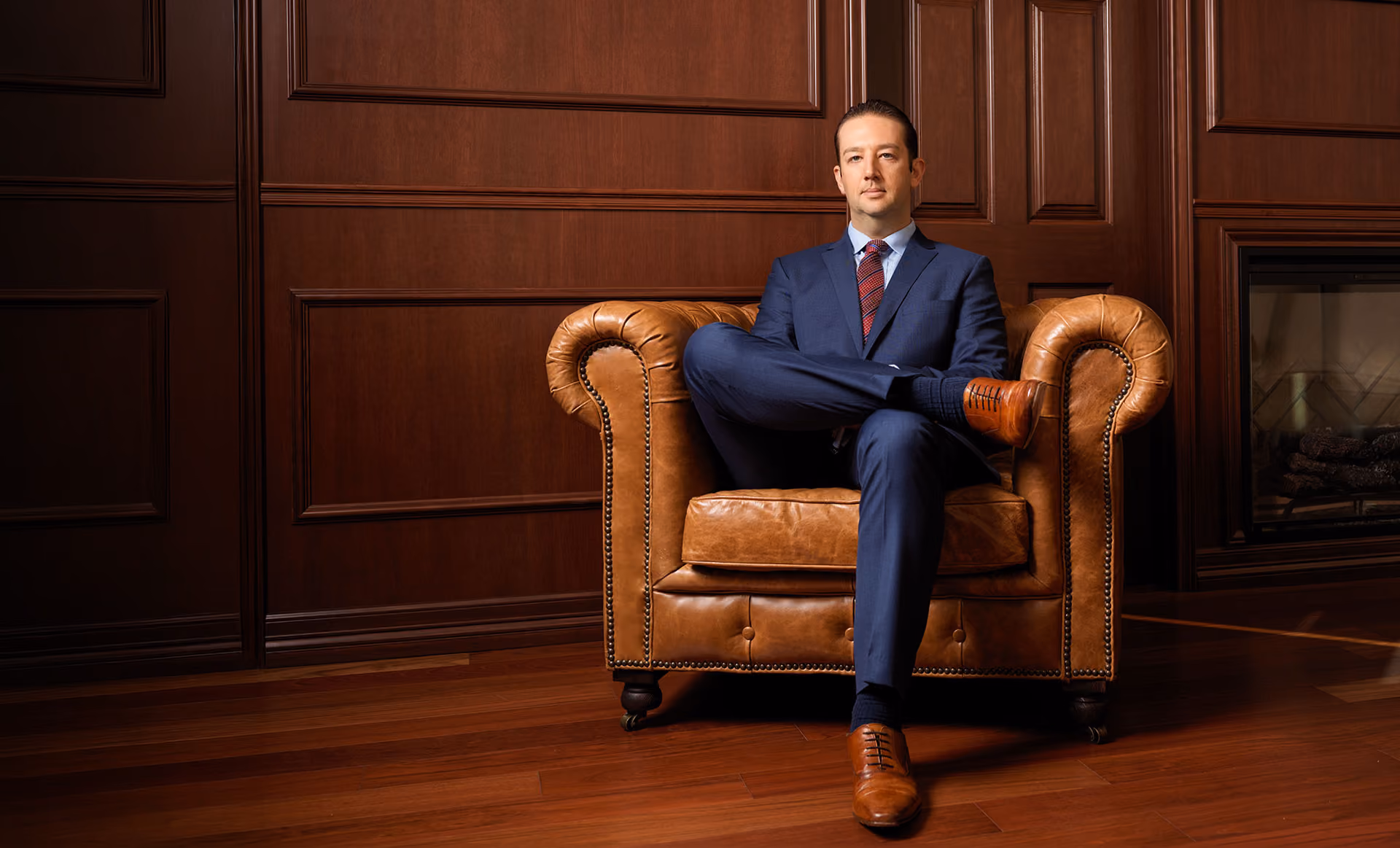 Man in a navy suit sitting cross-legged on a brown leather armchair in a wood-paneled room.