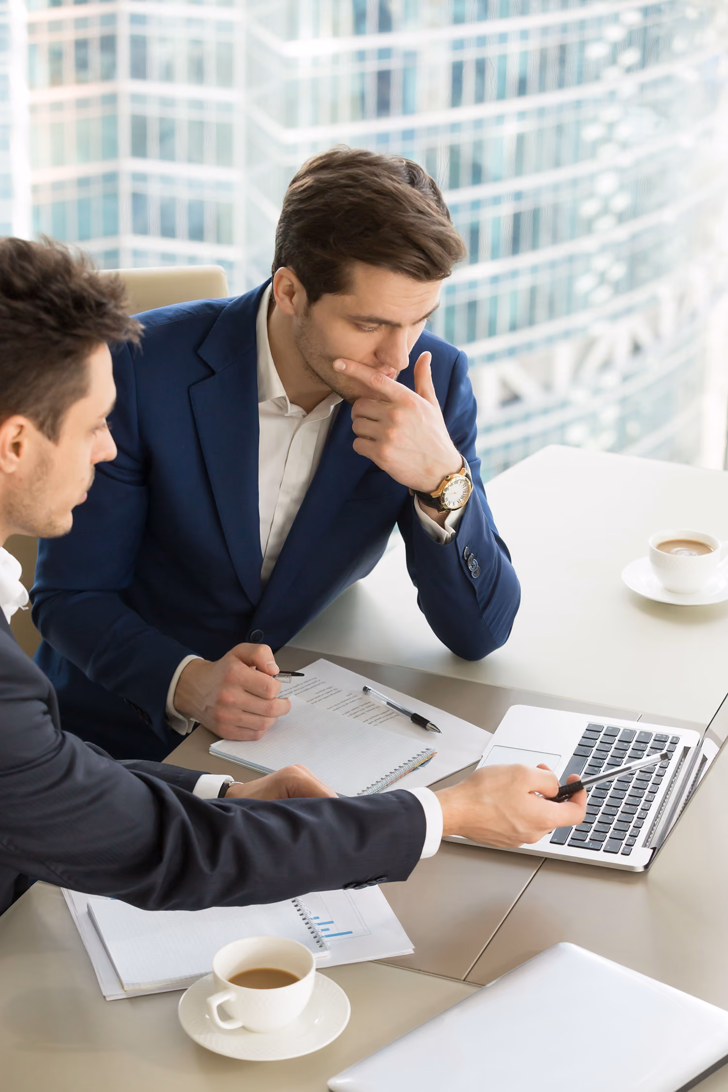 Two businessmen in suits discuss documents and laptop data over coffee in a modern office.