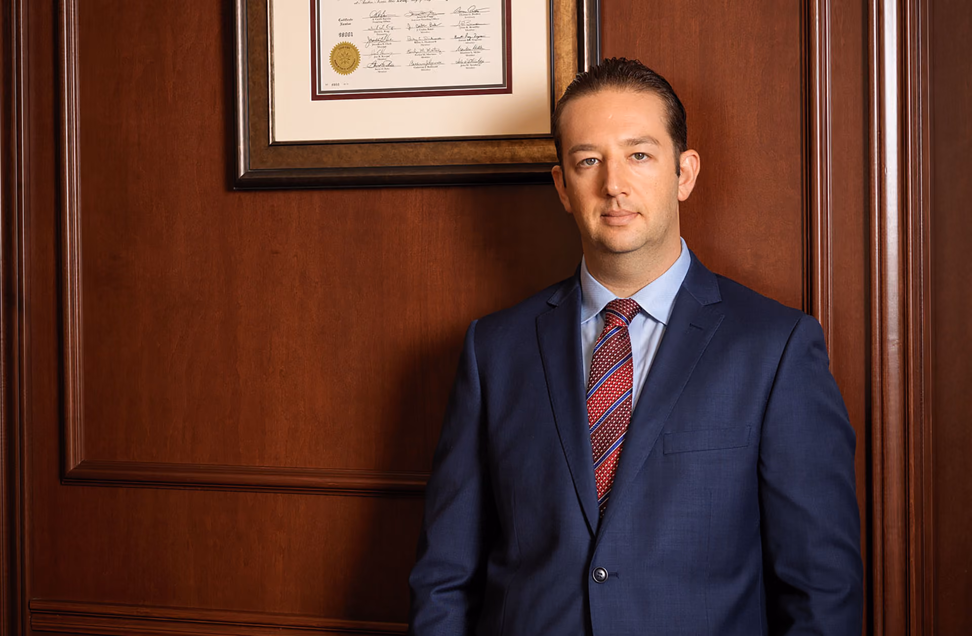 Man in blue suit and patterned tie standing in front of a wood-paneled wall with a framed certificate.