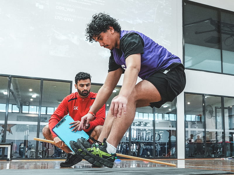 Athlete performing a long jump test while a SportsKey coach measures with a wooden stick