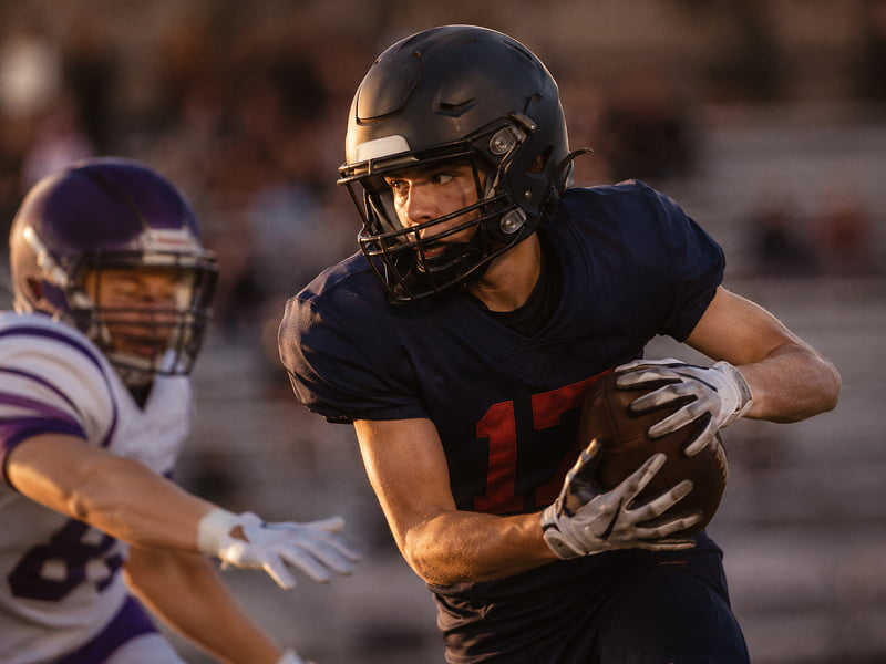 Focused football player in black helmet and jersey holding ball, avoiding opponent in white and purple uniform.