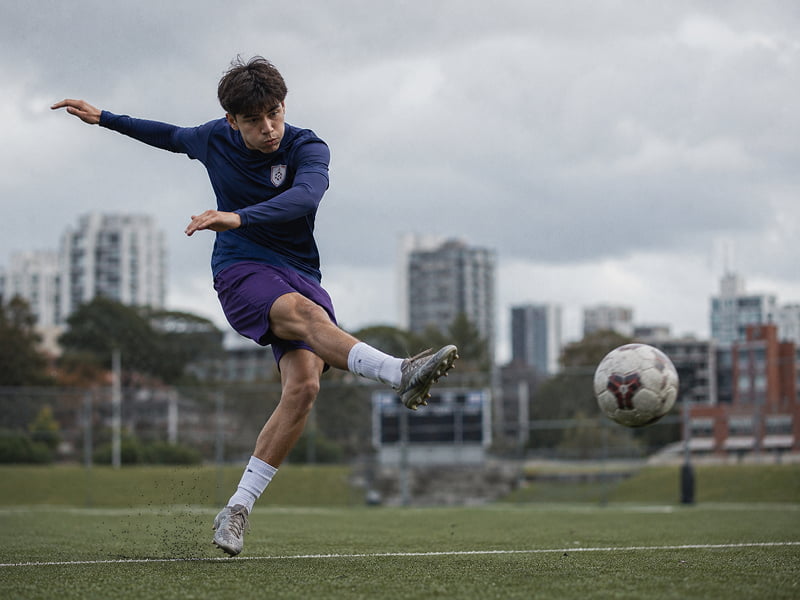 Young male soccer player kicking a ball on a grass field with city buildings in the background.