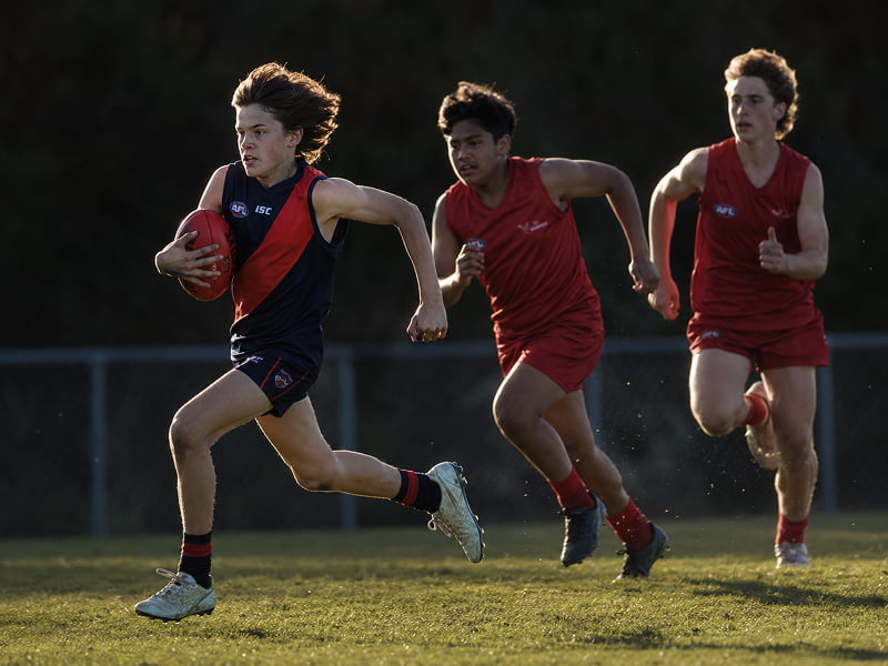 Young athlete in black and red runs with football while two opponents in red chase him on grass field.