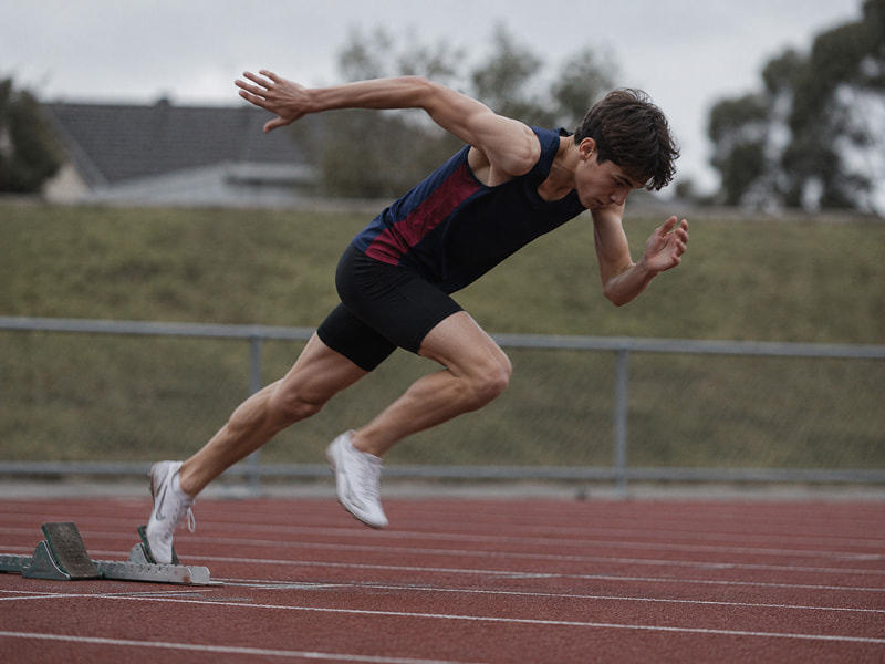 Athlete in black and navy gear sprinting off starting blocks on an outdoor track.