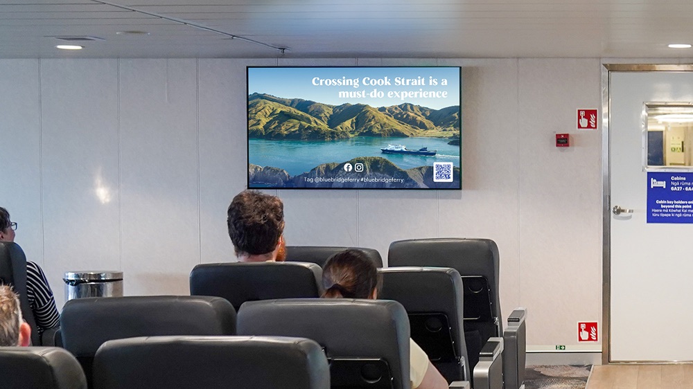 Interior of a ferry lounge with passengers seated facing a wall-mounted screen displaying an advertisement for Bluebridge Ferry, featuring a scenic photo of Cook Strait with a ferry on the water and the text 'Crossing Cook Strait is a must-do experience.'
