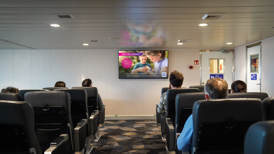 Passengers seated in rows of black chairs inside a ferry or boat cabin, facing a wall with a digital screen displaying an educational message about ladybugs and wildlife protection.