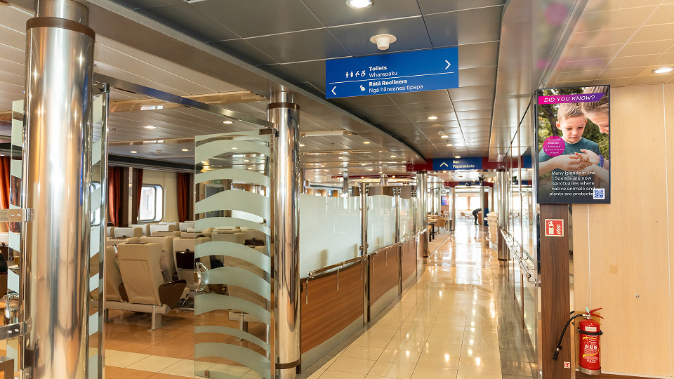 Interior of a ferry or ship showing a brightly lit corridor with polished floors, stainless steel columns, frosted glass partitions, and beige seats arranged in rows to the left, with blue overhead signs indicating directions to toilets and recliners.