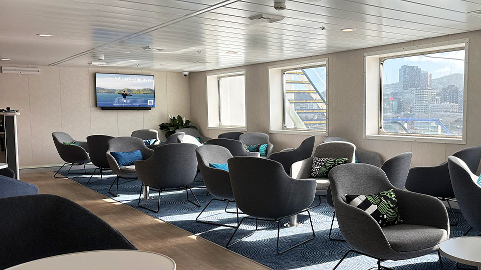 Interior of a modern lounge with gray armchairs arranged in pairs on a blue patterned carpet, small round white tables, large windows showing a cityscape, and a TV mounted on the wall displaying an outdoor scene.