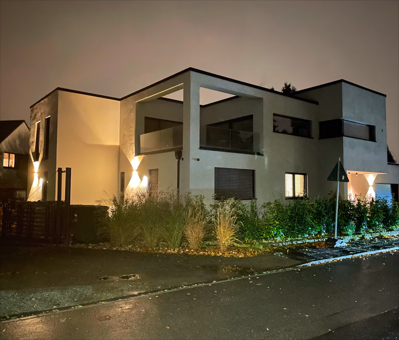 Modern two-story house illuminated by exterior lights on a wet street at night.
