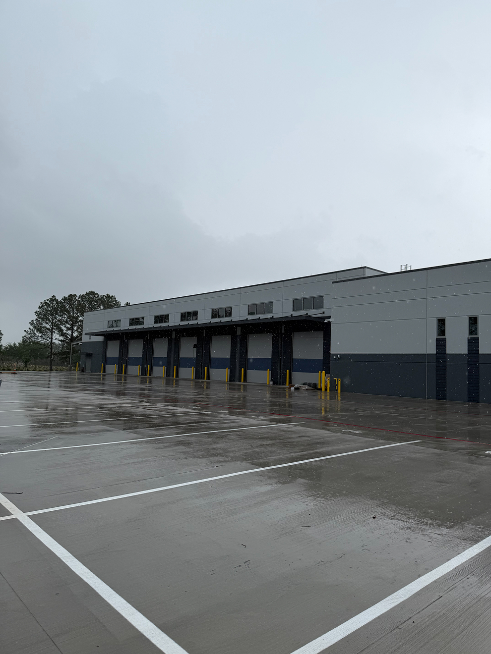 Wet parking lot in front of a modern warehouse building with closed loading bays on a cloudy day.