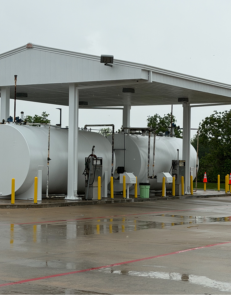 White industrial fuel tanks with pumps under a metal canopy on a wet concrete surface.