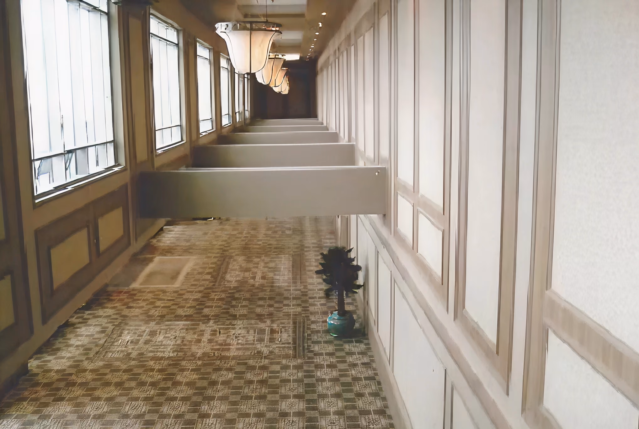 Empty corridor with tile floor, wall paneling, windows, hanging lamps, and a potted plant.