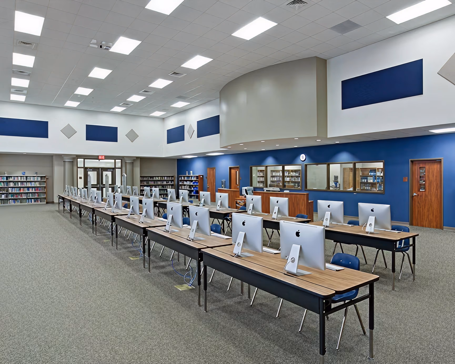 Library computer lab with rows of iMacs on tables facing away from the camera and bookshelves in the background.