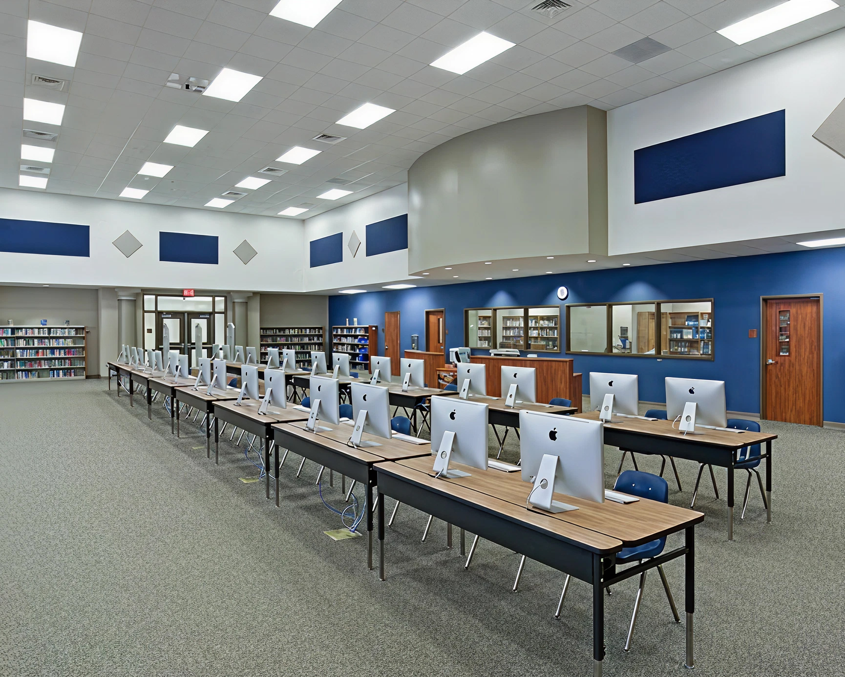 Library computer lab with rows of iMacs on tables facing away from the camera and bookshelves in the background.