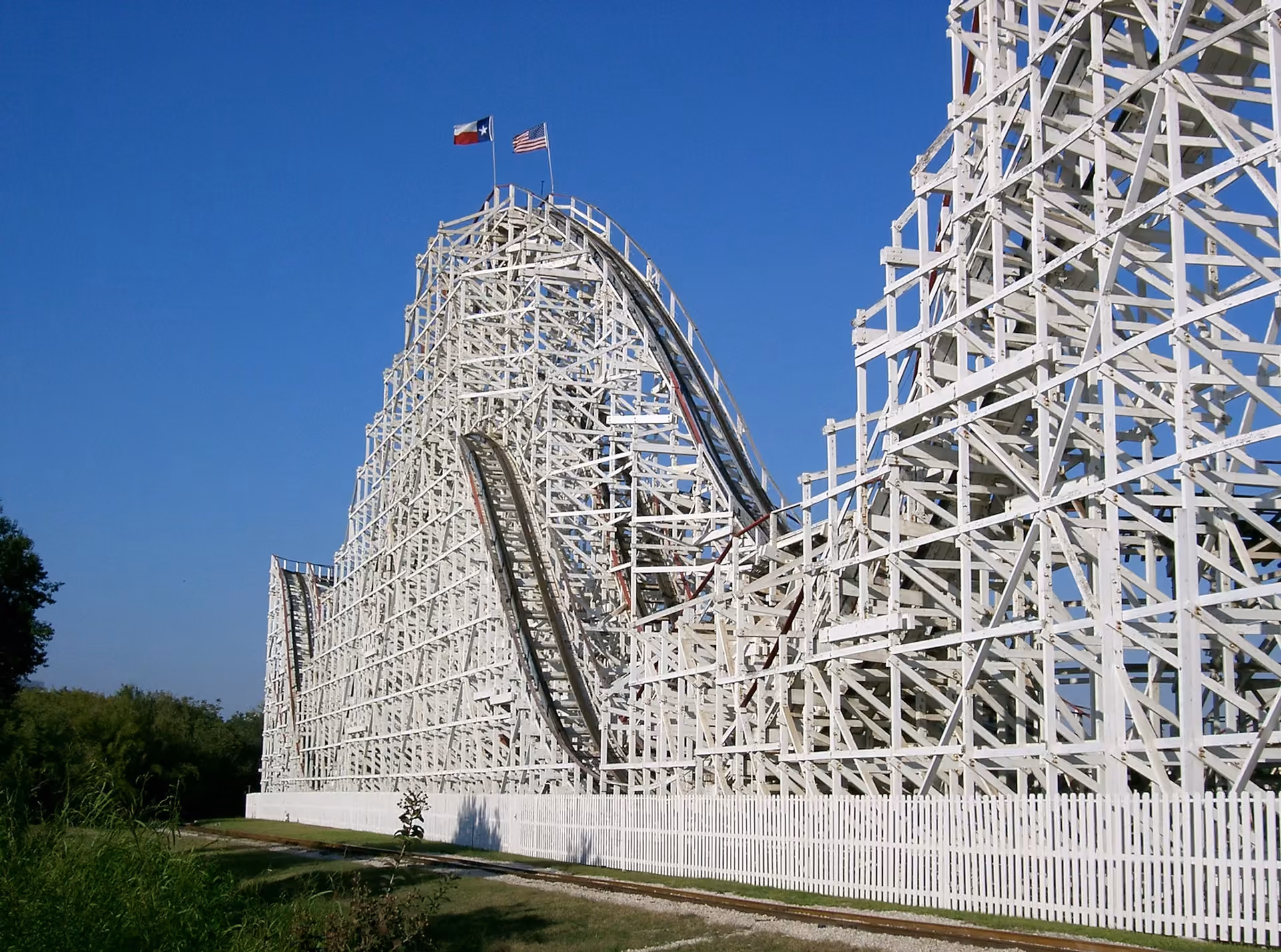 Large wooden roller coaster with Texas and American flags atop a peak against a clear blue sky.