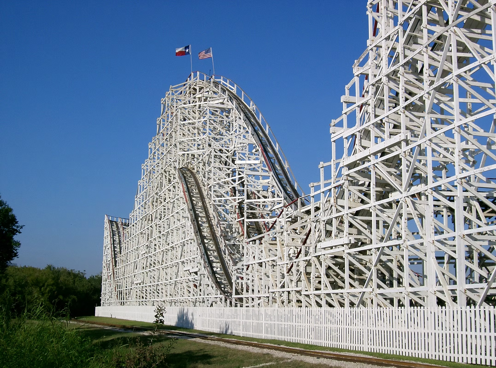 Large wooden roller coaster with Texas and American flags atop a peak against a clear blue sky.