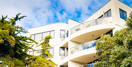 Modern white multi-story building with balconies surrounded by green trees under a blue sky.