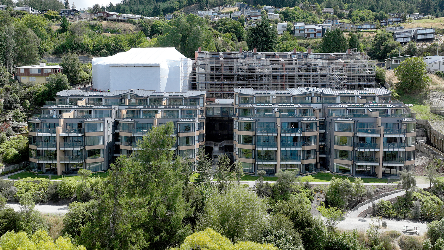 Modern apartment buildings with large glass windows surrounded by greenery and hills with houses above.
