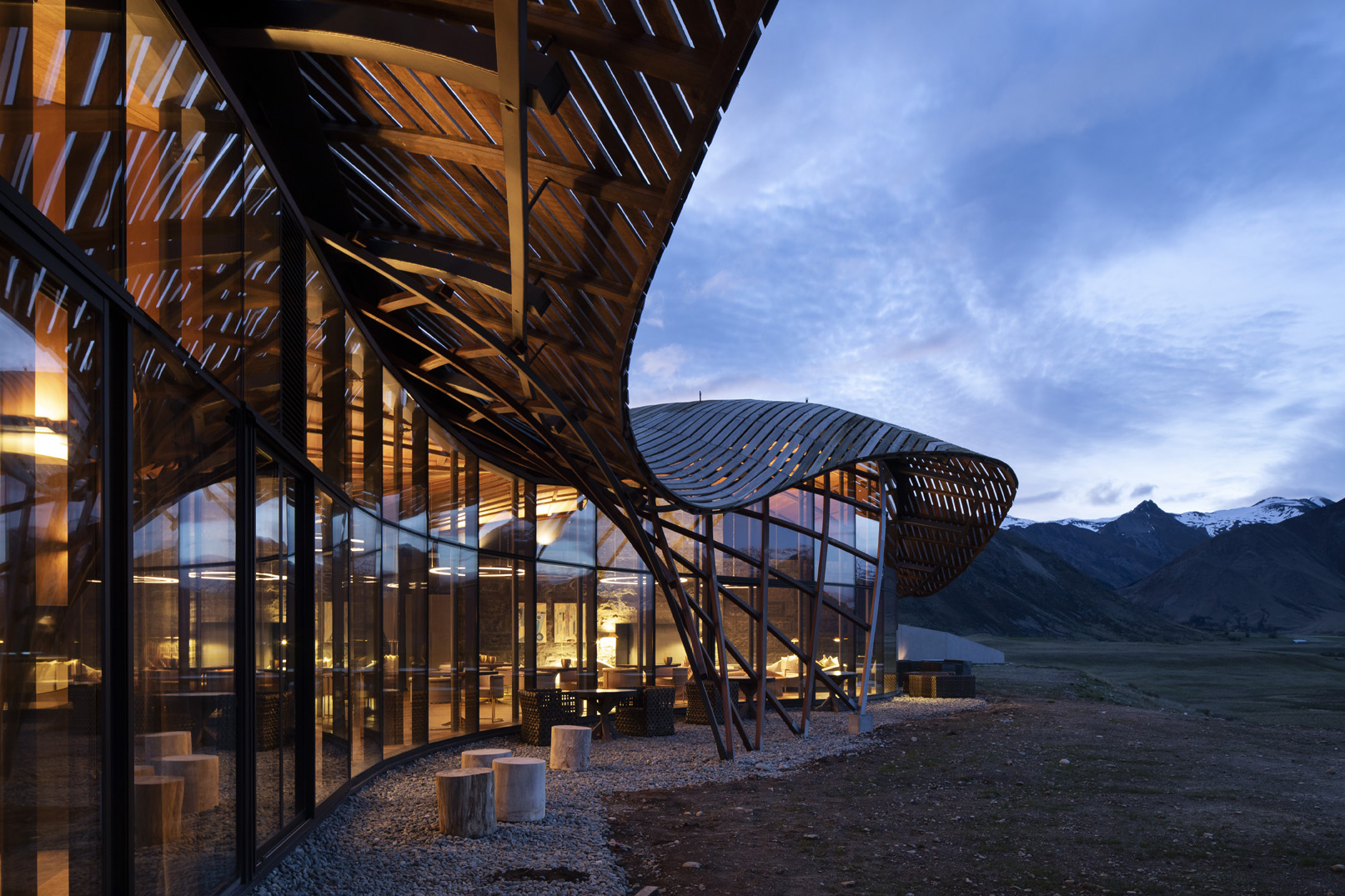 Curved wooden roof and glass walls with outdoor seating at dusk near mountains with snow.