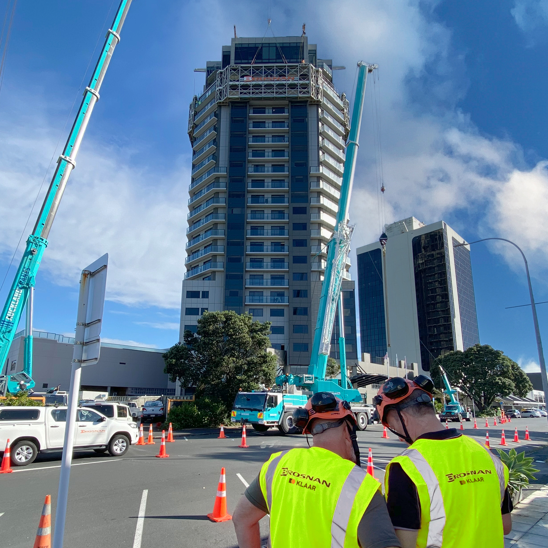 Two workers with Brosnan Klaar vests and helmets near cranes working on a tall building under blue sky.