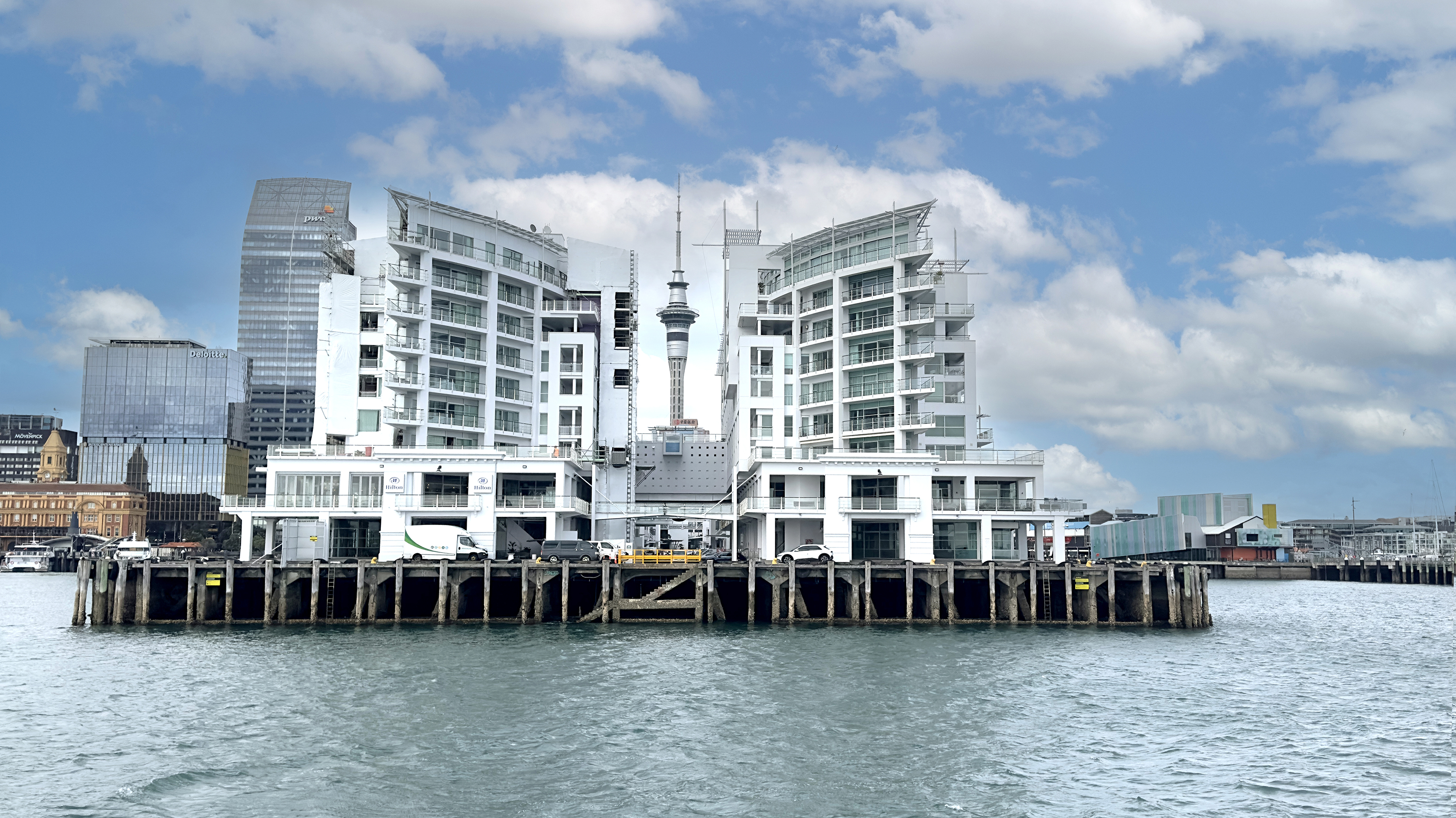 Harbor view of white modern buildings on pier with city skyline and tower under blue sky with clouds.
