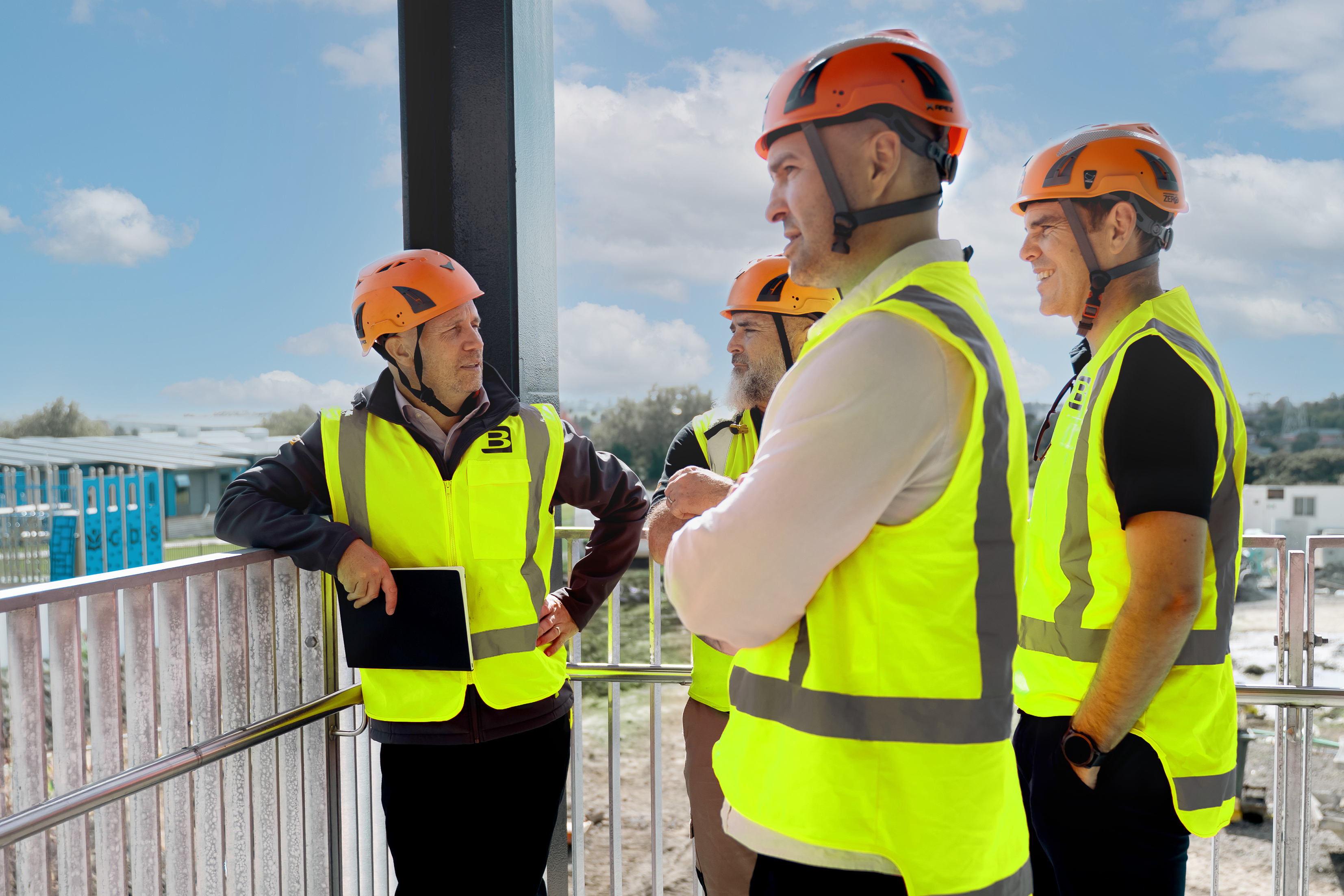 Four construction workers in orange helmets and yellow vests talking outdoors on a sunny day.